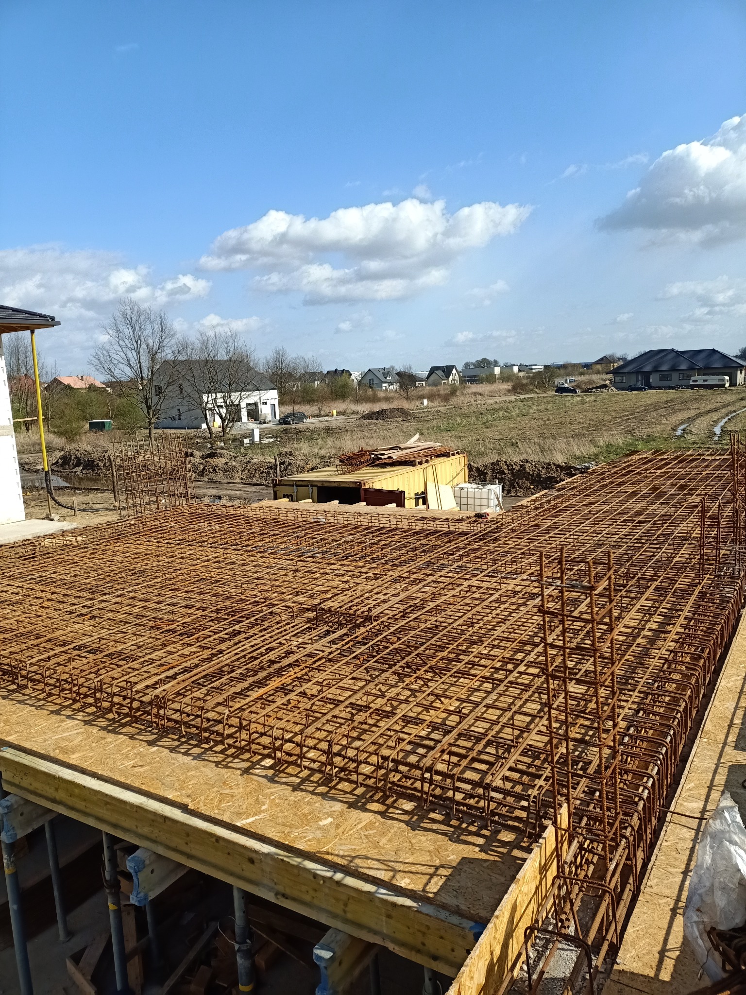 Siatka zbrojeniowa ułożona na deskowaniu, przygotowana do zalania betonem, widok z góry na plac budowy z oddalonymi zabudowaniami w tle, słoneczny dzień.