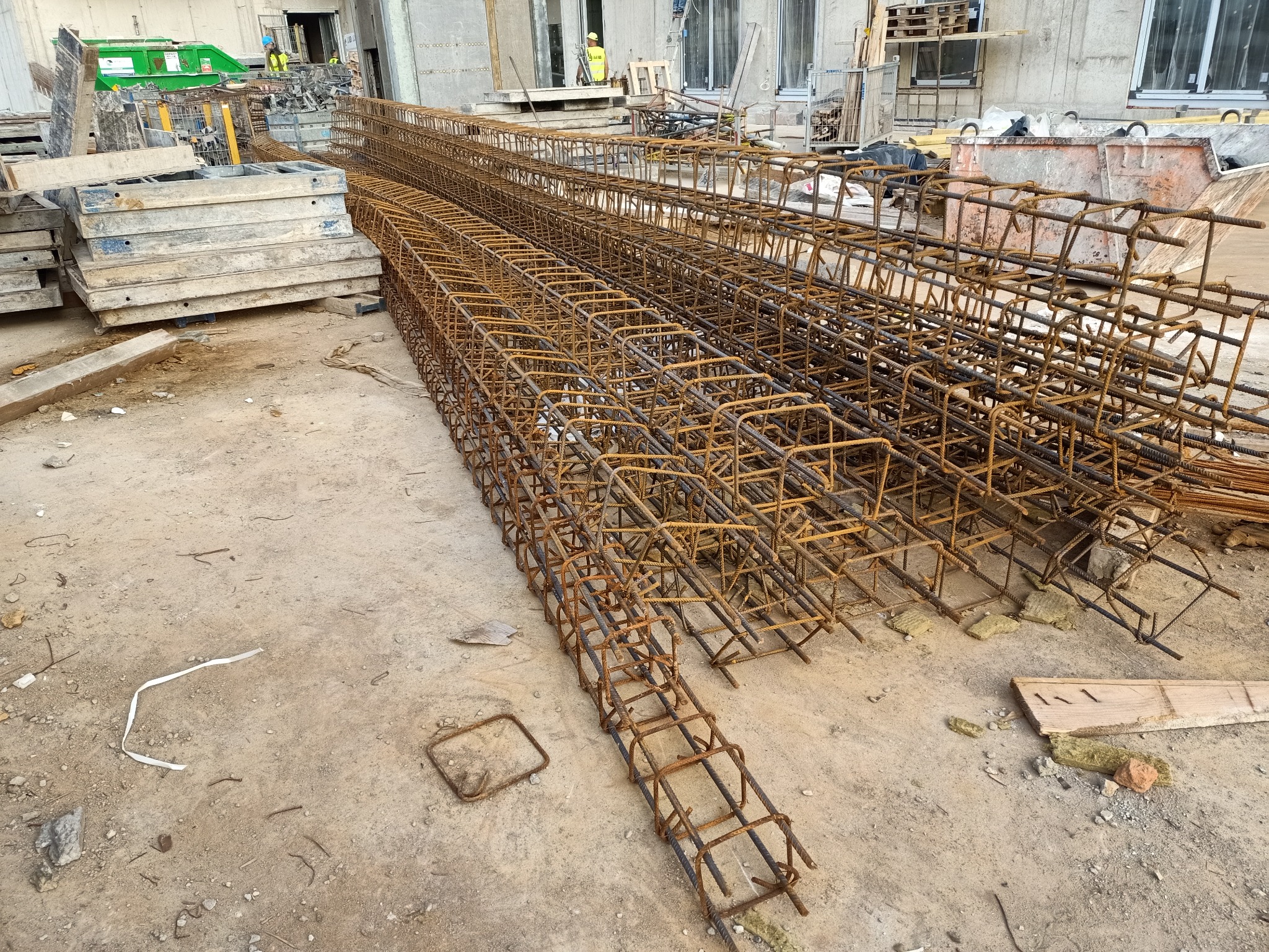 Spięte zbrojenie w formie klatek ułożone na placu budowy, widoczne szalunki i kontener w tle.