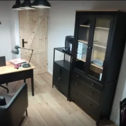 Jasne biuro z drewnianym biurkiem, czarnym krzesłem, regałem i lampami wiszącymi w minimalistycznym stylu.