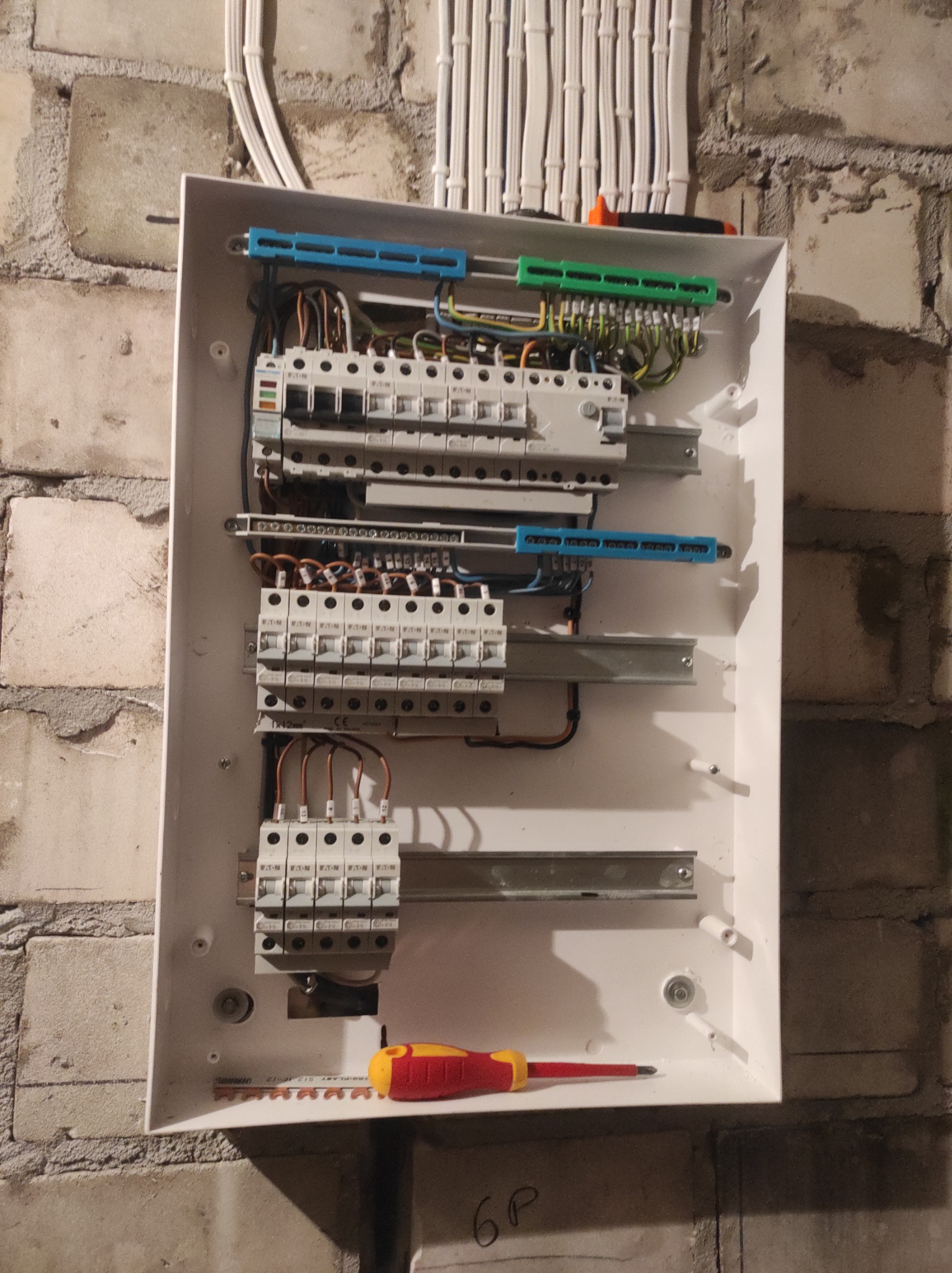 Otwarta skrzynka bezpiecznikowa z zamontowanymi bezpiecznikami i podłączonymi przewodami, zamontowana na surowej, ceglanej ścianie, z widocznymi narzędziami: wkrętakiem z czerwoną i żółtą...