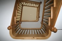 Spiralne schody drewniane z jasnego drewna, widok z dołu, ukazujący zakrzywioną poręcz z okrągłymi zakończeniami i stopniowane stopnie w perspektywie.