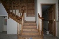 Drewniane schody z balustradą i poręczą w domu w Leszczach, widok z dołu, z otwartymi drzwiami w tle.