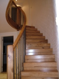 Kręte schody z jasnego drewna z metalowymi prętami balustrady, widok z dołu, na ścianie dekoracyjna faktura.