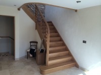 Klatka schodowa z drewnianymi schodami o łagodnym, zakrzywionym biegu, z ażurową balustradą i jasnymi ścianami. Na podeście stoi krzesło i skrzynka.