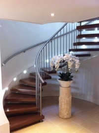 Kręte schody z drewnianymi stopniami i metalową poręczą, oświetlone punktowo, z dekoracyjną kolumną i kwiatami na podeście.