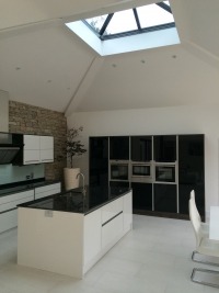 Nowoczesna kuchnia z wyspą, czarno-białe szafki na wysoki połysk, kamienna ściana i doświetlenie przez okno dachowe.