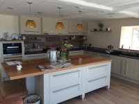 Jasna kuchnia z wyspą i drewnianym blatem, kremowe szafki z metalowymi uchwytami, nowoczesne oświetlenie wiszące, widoczne elementy dekoracyjne i sprzęt AGD.