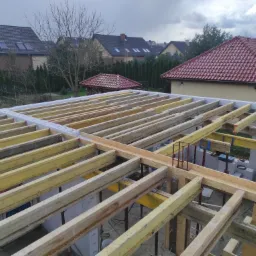 Surowa konstrukcja stropu z drewnianych belek i białych bloczków, widok z góry na budowę domu jednorodzinnego w pochmurny dzień.