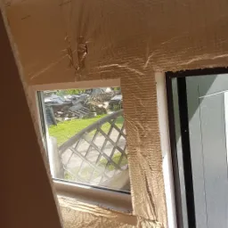 Izolacja termiczna folią aluminiową wok&oacute;ł nowo wstawionego okna z widokiem na zagracony ogr&oacute;d.