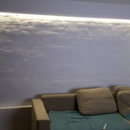 Ściana z dekoracyjną strukturą podświetloną listwą LED, szara sofa z poduszkami i turkusowym pledem w pokoju.