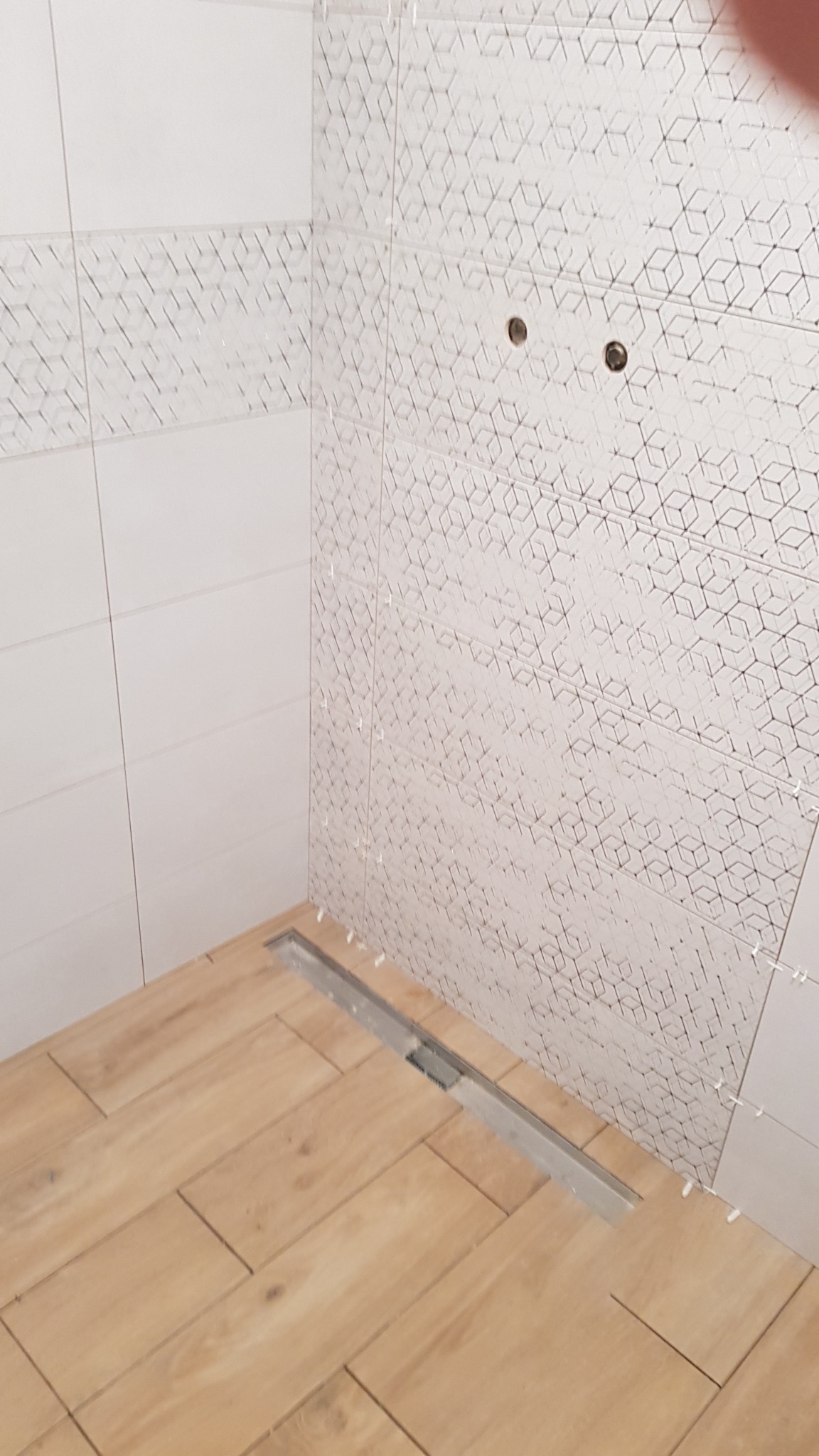 Układanie płytek podłogowych imitujących drewno w kabinie prysznicowej z odpływem liniowym, ściany wyłożone białymi płytkami z geometrycznym wzorem, widoczne krzyżyki dystansowe.