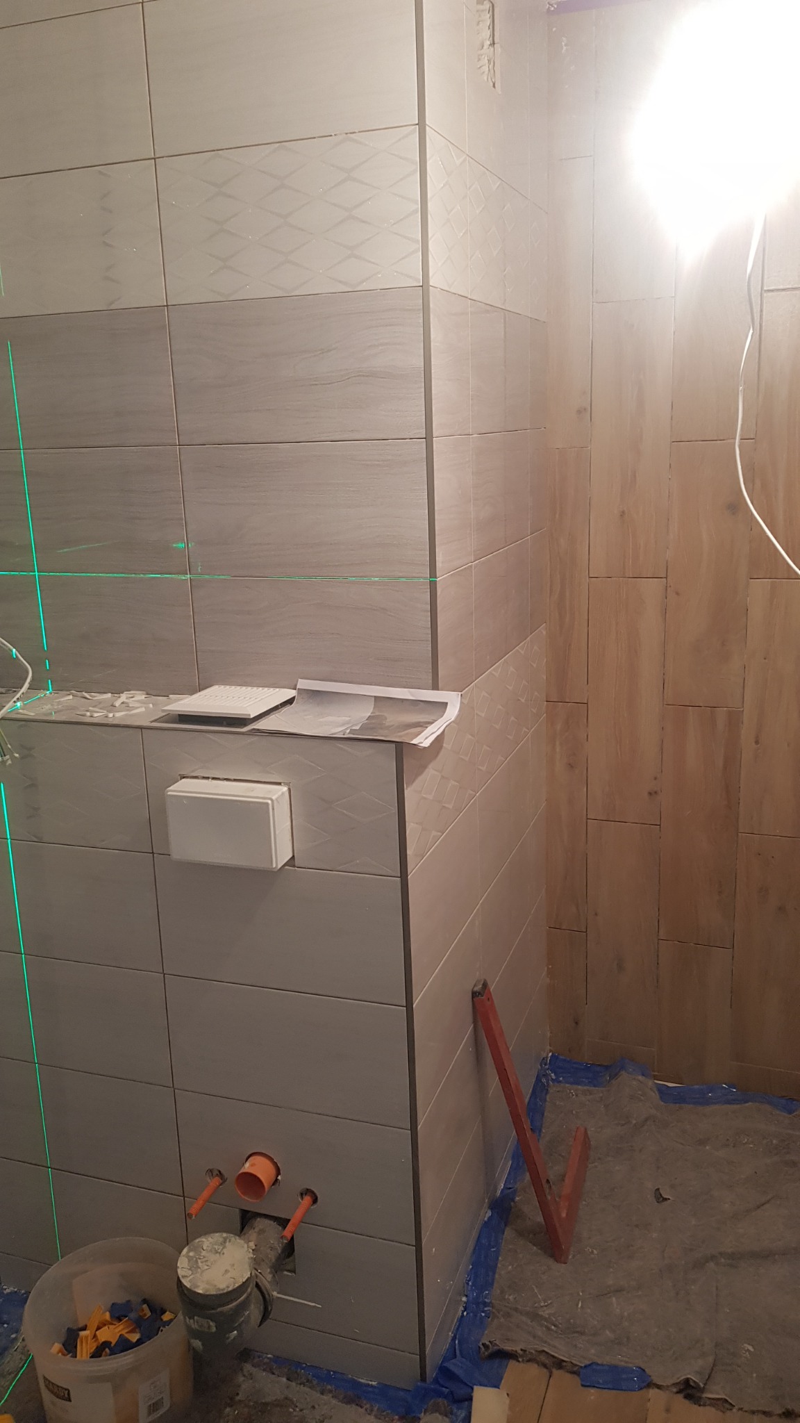 Ściana łazienki w trakcie remontu: częściowo wyłożona szarymi płytkami z delikatnym wzorem, widoczny laserowy poziom, rury instalacyjne wystające ze ściany, kubeł z akcesoriami i narzędzia...