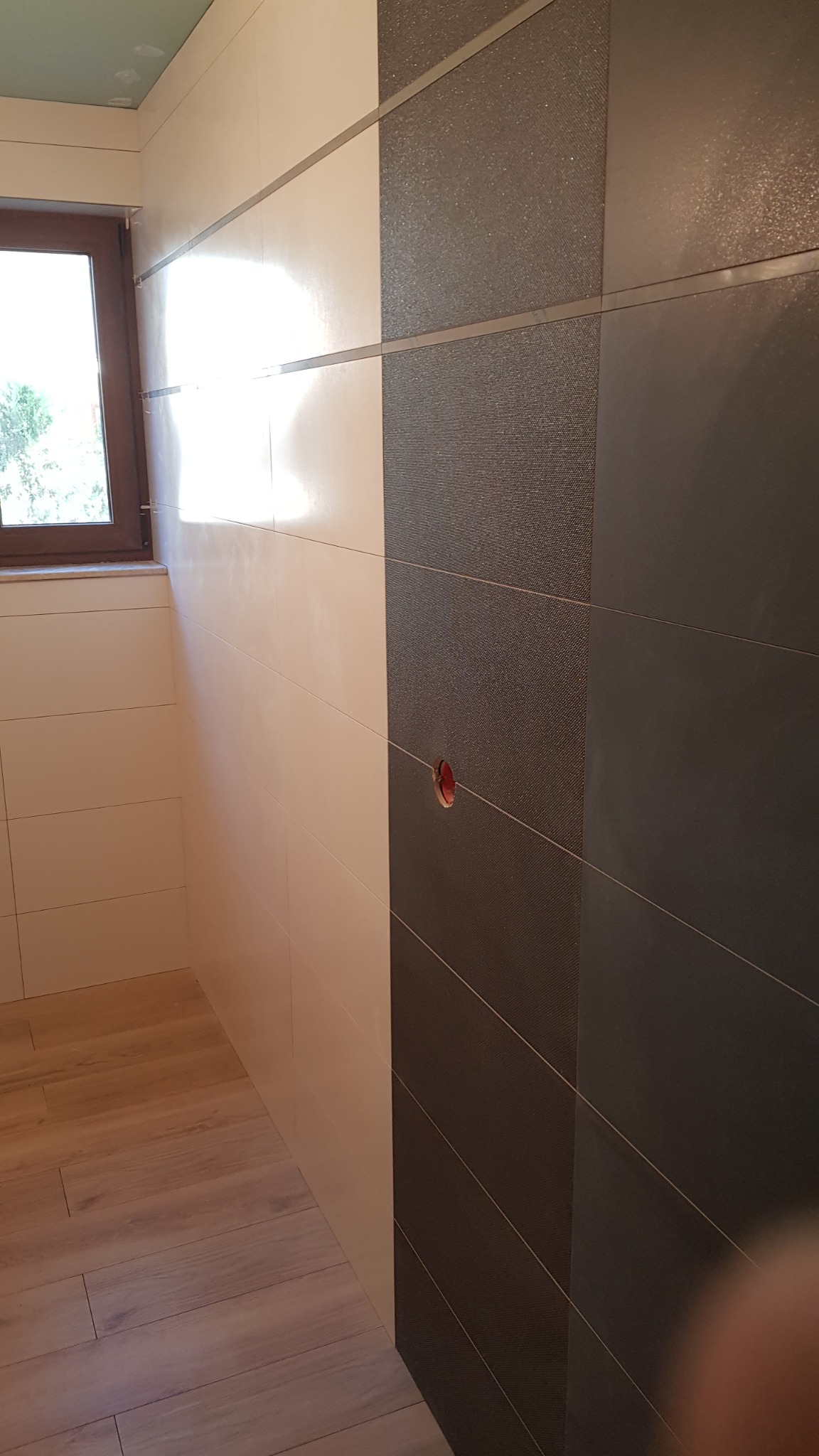 Fragment łazienki z wykończonymi ścianami: białe, błyszczące płytki ceramiczne zestawione z ciemnoszarymi płytkami o fakturze tkaniny, widoczne gniazdko elektryczne bez osprzętu, drewnopodobna...