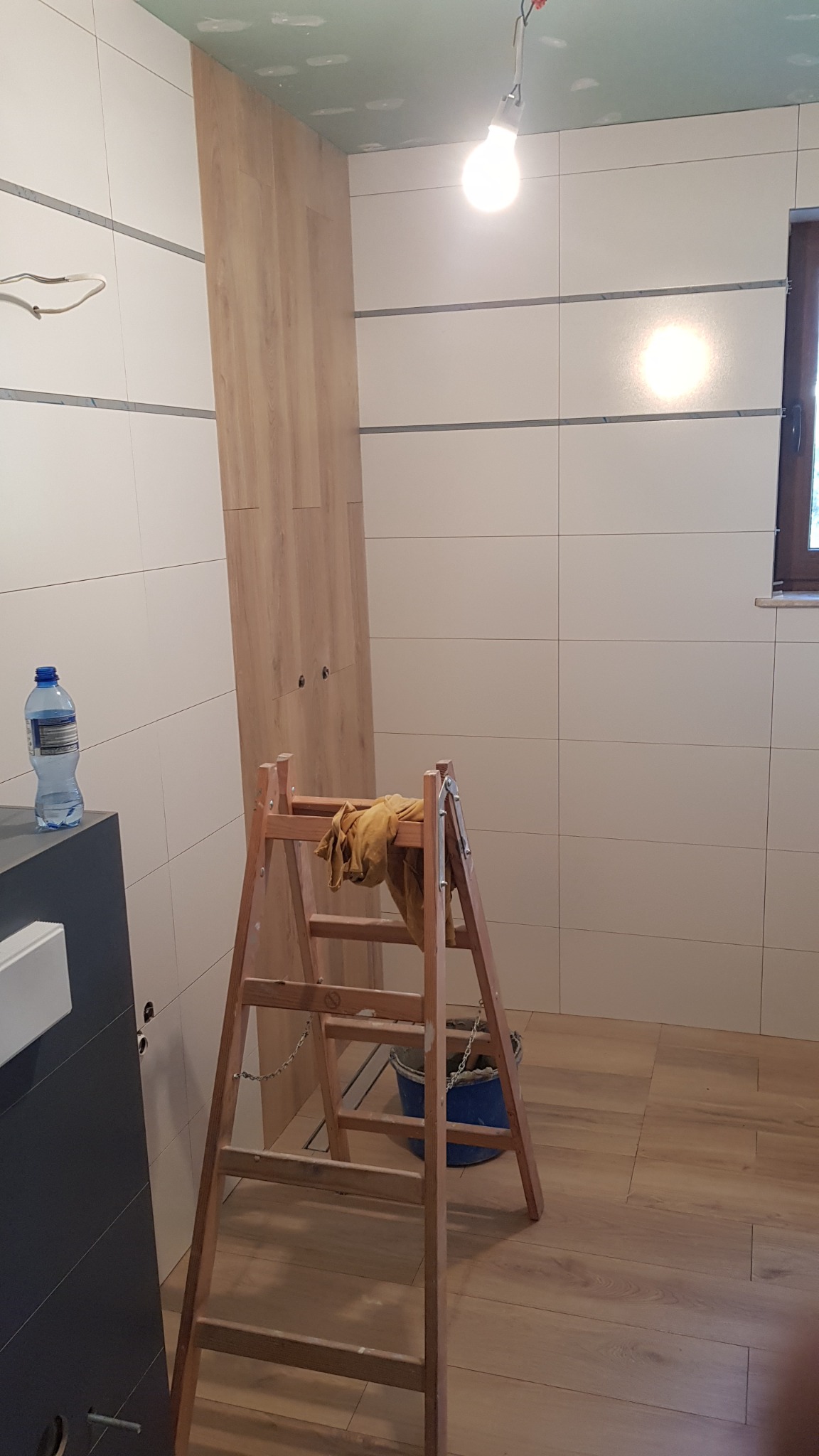 Wnętrze łazienki w trakcie prac: ułożone płytki ścienne i podłogowe imitujące drewno, drabina z wiadrem i szmatką, częściowo zamontowana szafka z frontami w kolorze drewna, sufit w trakcie...