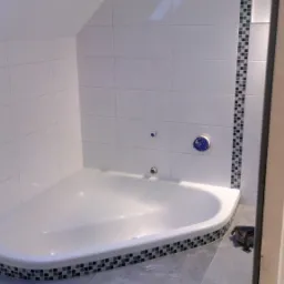 Biała narożna wanna z mozaikowym wykończeniem w łazience na poddaszu, z szarymi płytkami podłogowymi i dekoracyjnym pasem mozaiki na ścianie.