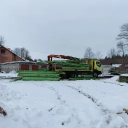 Żółta ciężarówka z dźwigiem rozładowuje impregnowane na zielono deski na zaśnieżonym placu, w tle budynek i drzewa bez liści.