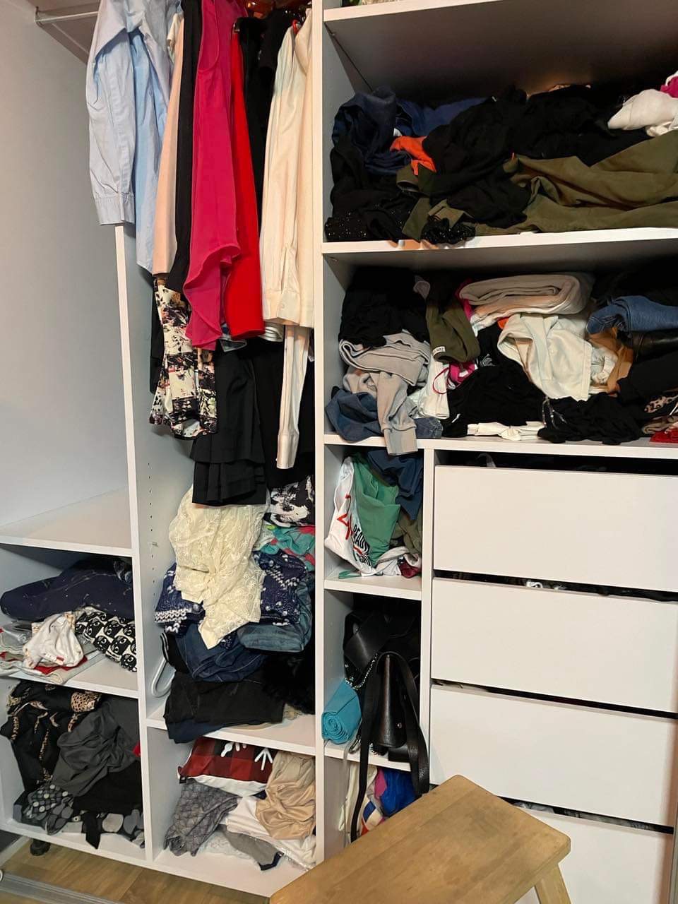 Niezorganizowana zawartość szafy z ubraniami, z półkami przepełnionymi różnorodnymi tkaninami i akcesoriami, obok białych szuflad i drewnianego stołka.