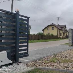 Nowoczesna, przesuwna brama w kolorze grafitowym z zamontowanym napędem Nice Robus, widoczna na tle domu jednorodzinnego i brukowanej drogi dojazdowej.