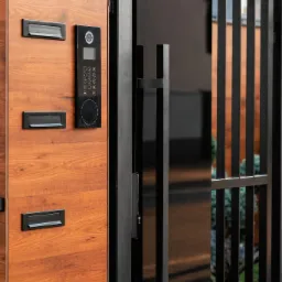 Nowoczesna brama w Siedlcach z drewnopodobną skrzynką na listy i panelem domofonu z klawiaturą numeryczną, połączona z czarnym, metalowym ogrodzeniem.