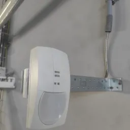 Biały czujnik ruchu zamontowany na metalowej listwie z otworami, przymocowanej do szarej ściany. Nad czujnikiem widoczna szara rurka instalacyjna. W tle widoczne koryto kablowe z przewodami.