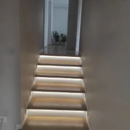 Nowoczesne schody drewniane z podświetleniem LED na każdym stopniu, prowadzące do jasnego pomieszczenia z roślinami w tle.