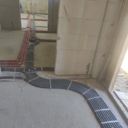 Multiwersum Energii Grzymała Michał - Wiązka szarych, karbowanych rur instalacyjnych przymocowana metalowymi obejmami do surowej podłogi w trakcie prac wykończeniowych, z widocznymi czerwonymi rurami na ścianach z betonu komórkowego.