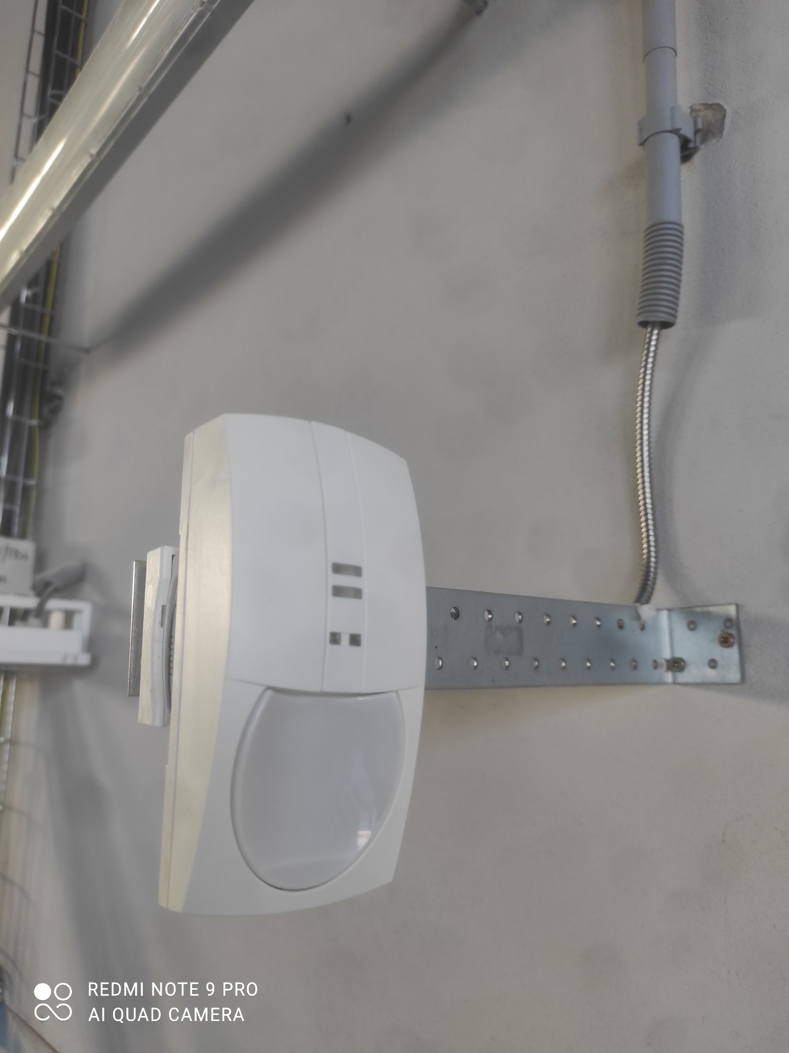 Biały czujnik ruchu zamontowany na metalowej listwie z otworami, przymocowanej do szarej ściany. Nad czujnikiem widoczna szara rurka instalacyjna. W tle widoczne koryto kablowe z przewodami.