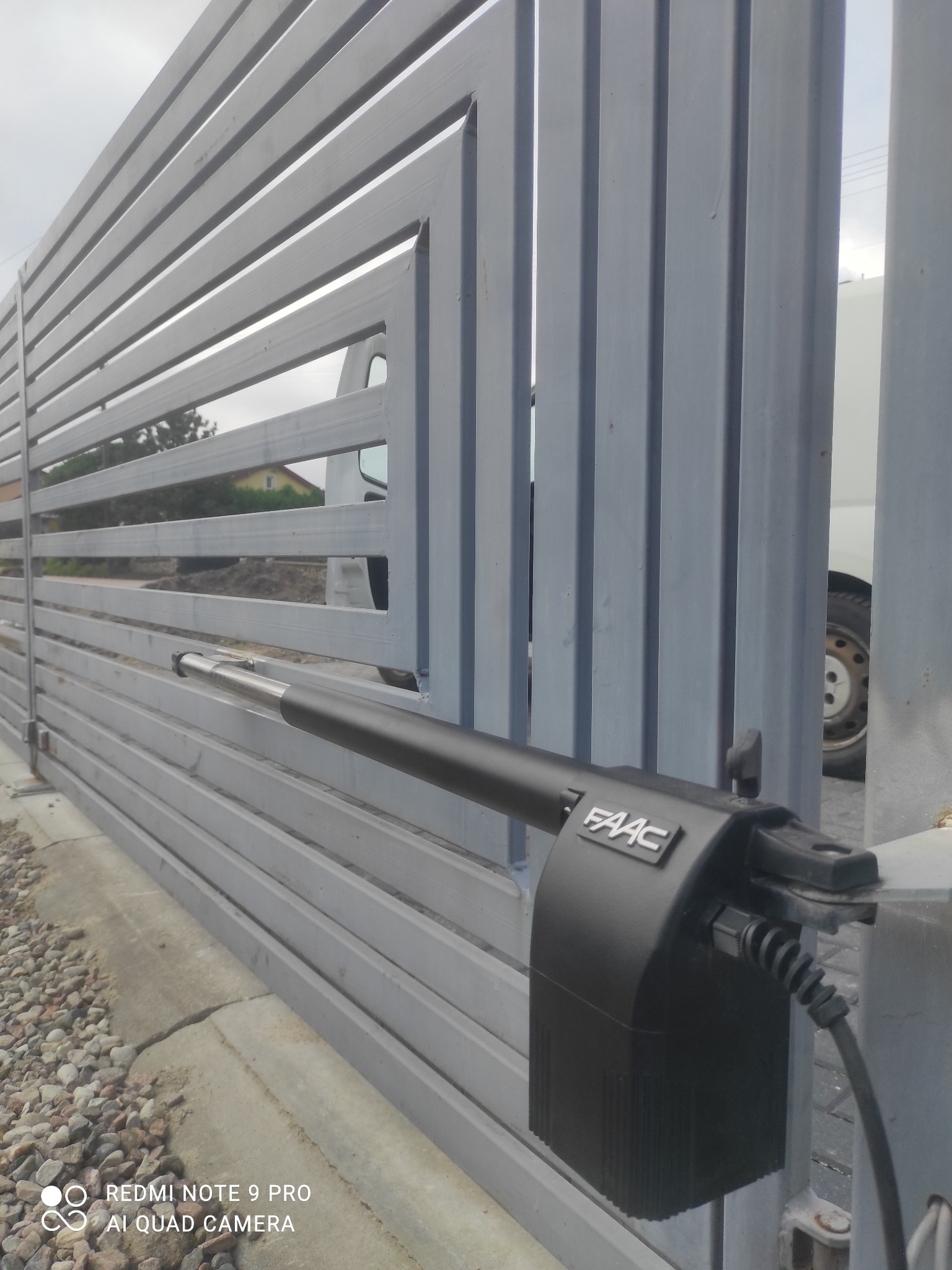 Szara, pozioma brama wjazdowa z zamontowanym czarnym siłownikiem FAAC; widoczny fragment białego samochodu w tle.