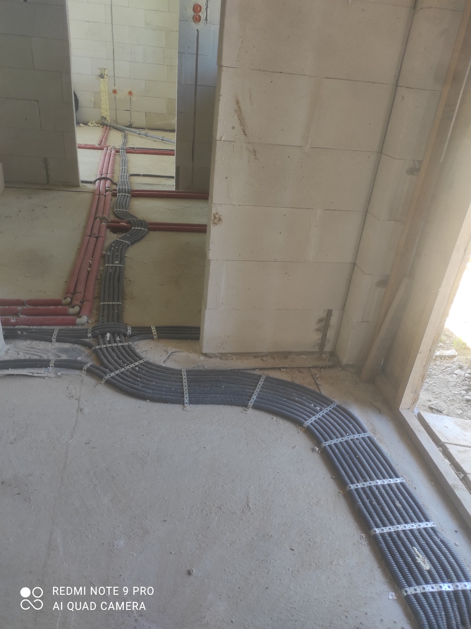 Wiązka szarych, karbowanych rur instalacyjnych przymocowana metalowymi obejmami do surowej podłogi w trakcie prac wykończeniowych, z widocznymi czerwonymi rurami na ścianach z betonu komórkowego.