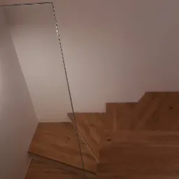 Nowoczesne schody z jasnego drewna z szklaną balustradą, oświetlone prostokątną lampą ścienną.