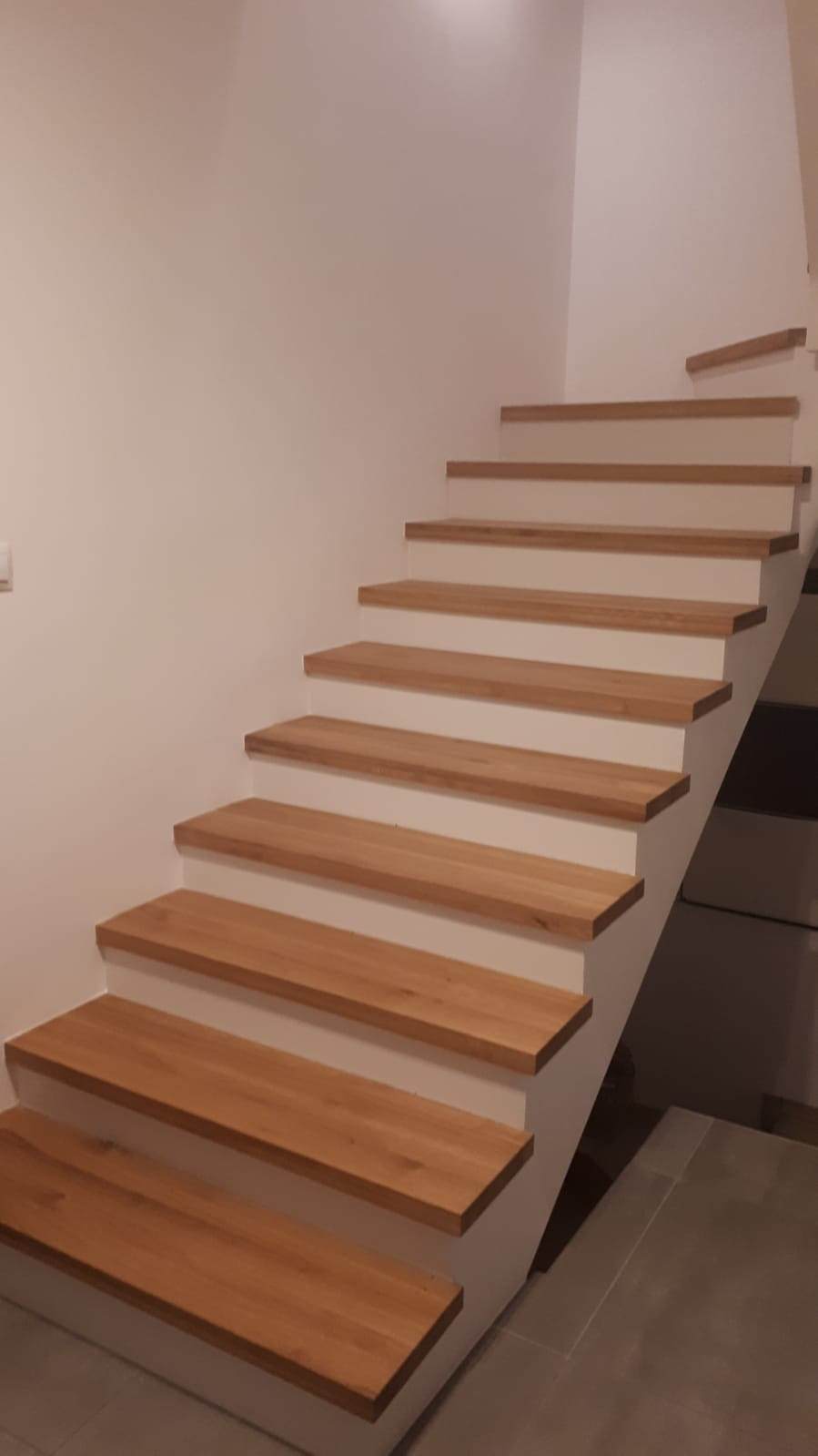 Proste, nowoczesne schody z drewnianymi stopniami i białymi podstopnicami, widoczne z dołu, z fragmentem szarej podłogi w tle.
