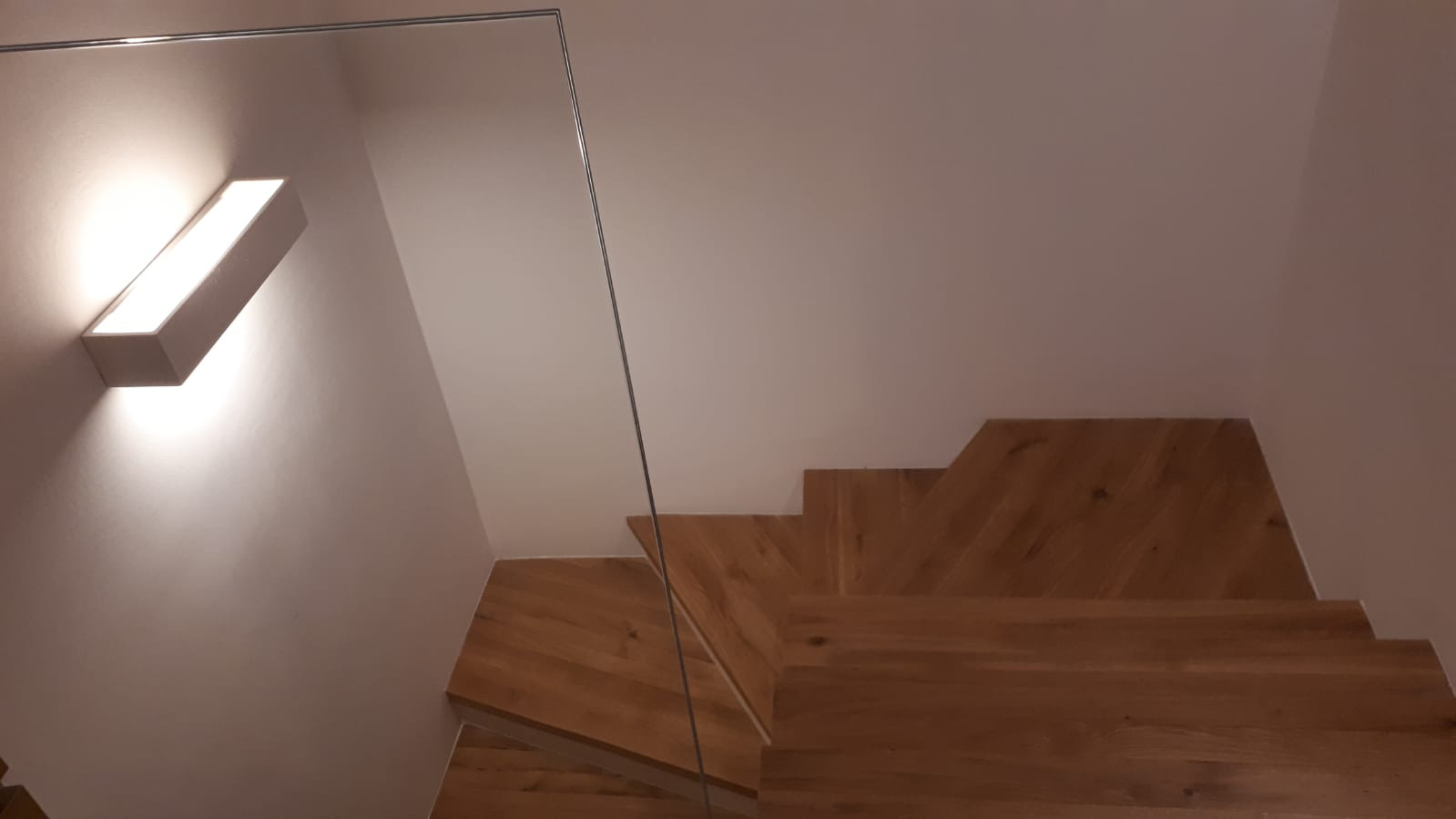 Nowoczesne schody z jasnego drewna z szklaną balustradą, oświetlone prostokątną lampą ścienną.