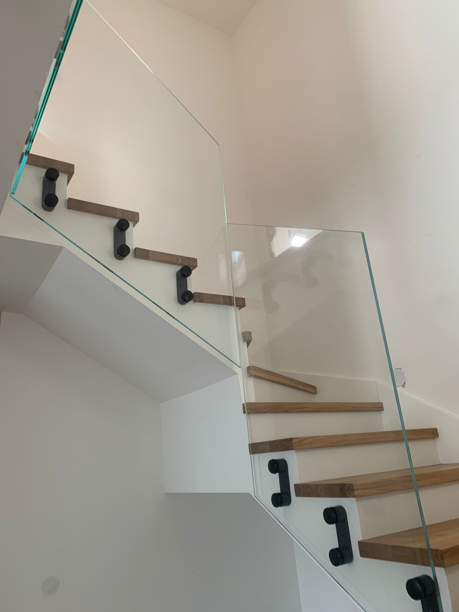 Nowoczesne schody z drewnianymi stopniami i szklaną balustradą, mocowaną czarnymi uchwytami do boku stopni.