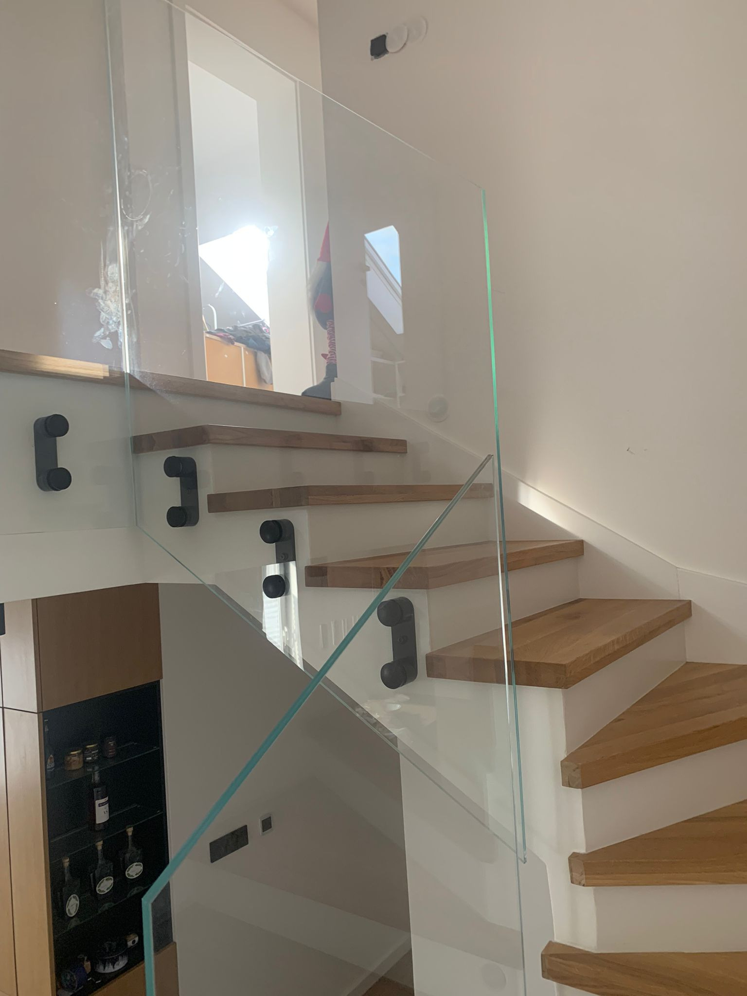 Drewniane schody z jasnymi stopniami i szklaną balustradą, mocowaną czarnymi, minimalistycznymi uchwytami, widok z dołu, z fragmentem zabudowy meblowej w tle.