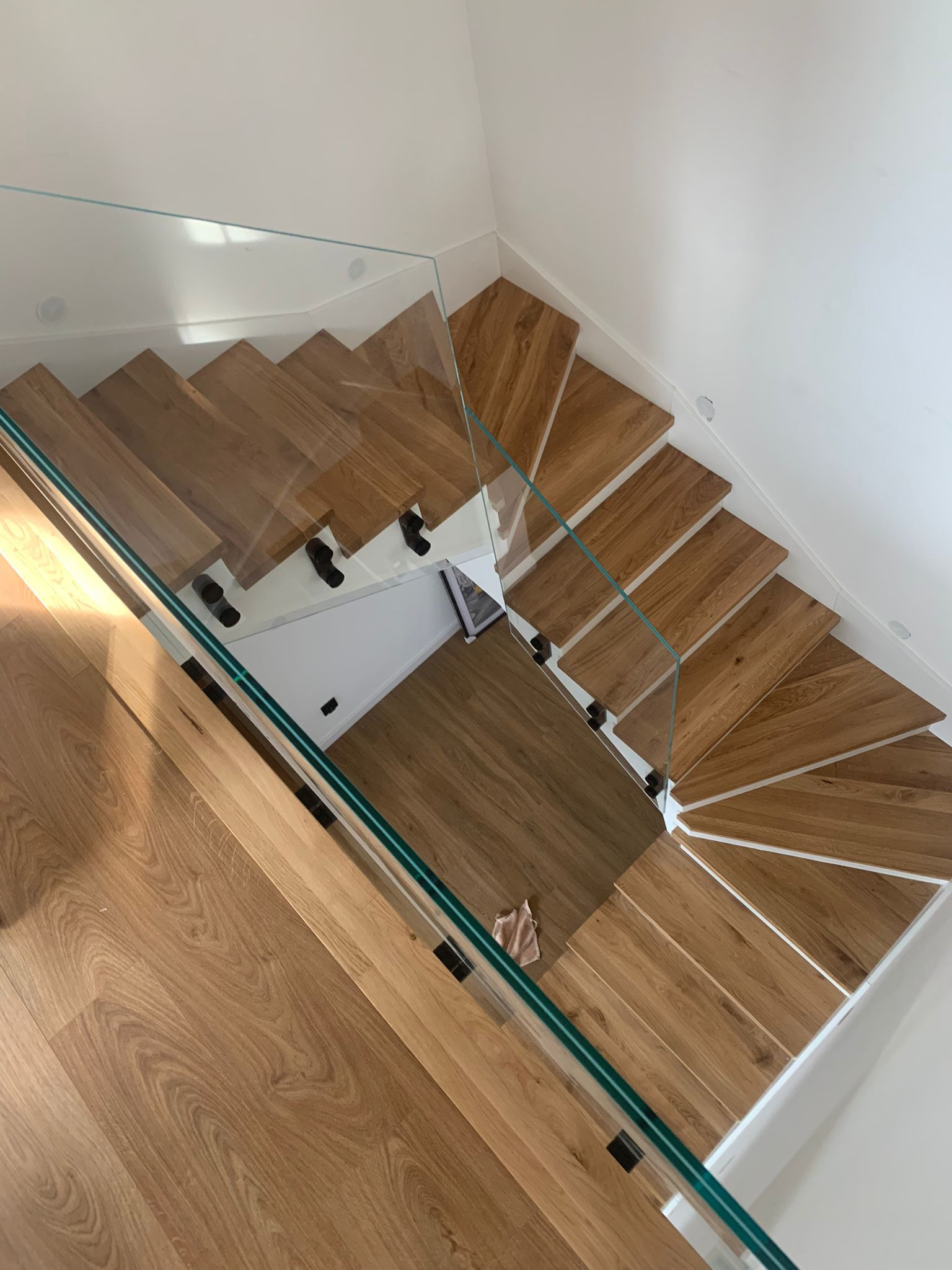 Kaskadowe schody z drewna dębowego z przezroczystą szklaną balustradą, widok z góry ukazuje naturalny rysunek słojów drewna i minimalistyczne czarne mocowania.