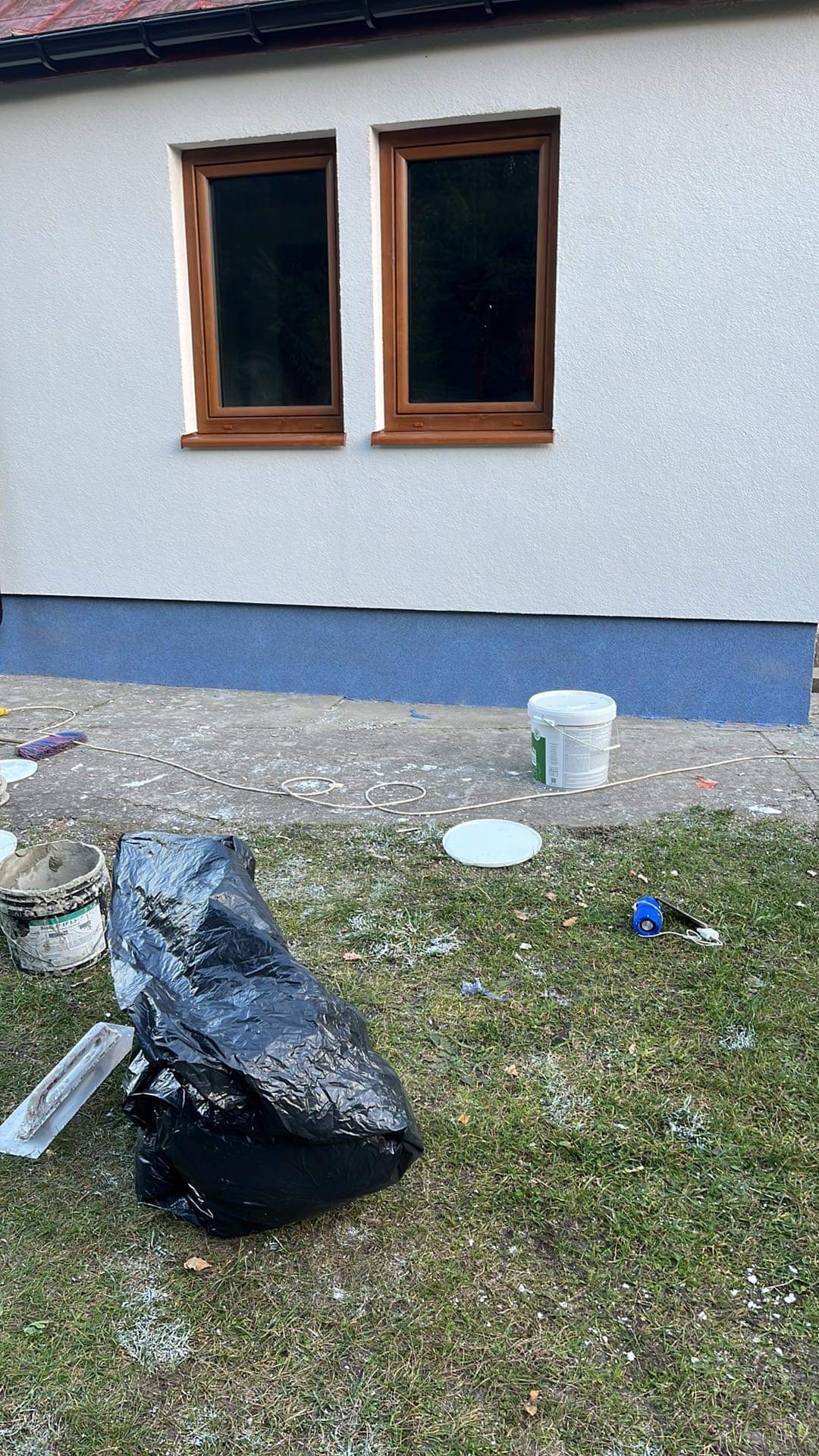 Świeżo pomalowana elewacja domu z brązowymi oknami, widoczne ślady farby na trawie, wiadro z farbą, pędzel i zakryta folią budowlaną zawartość.