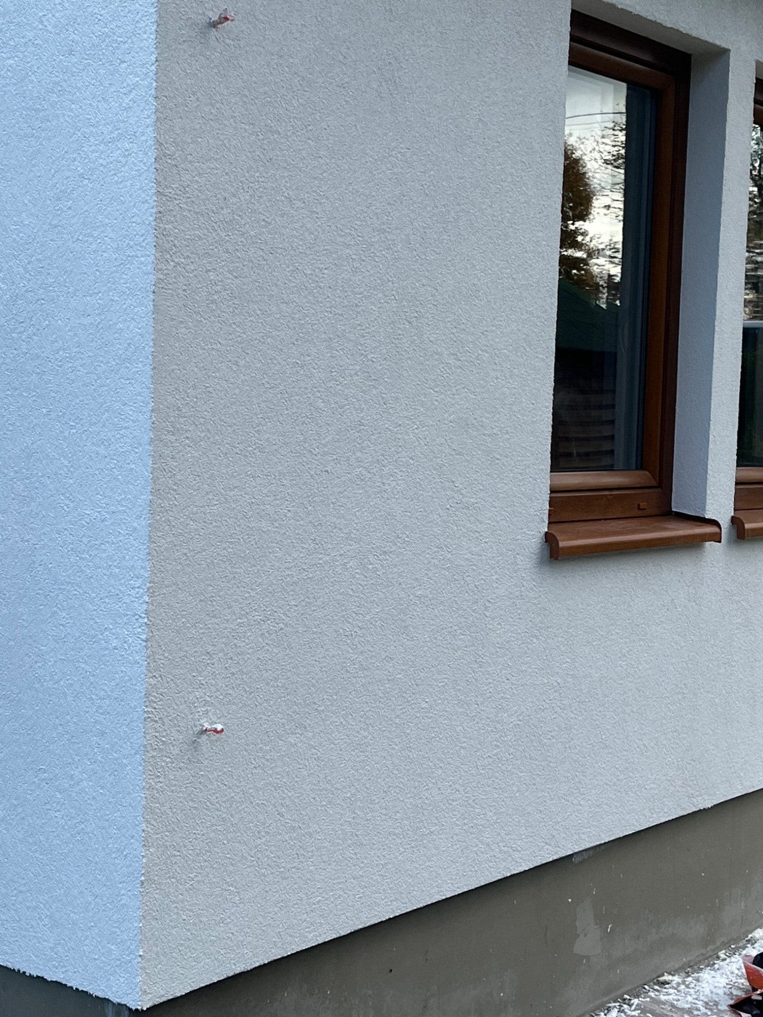 Fragment elewacji budynku z oknem w brązowej ramie, widoczne kołki montażowe na styku dwóch kolorów tynku.