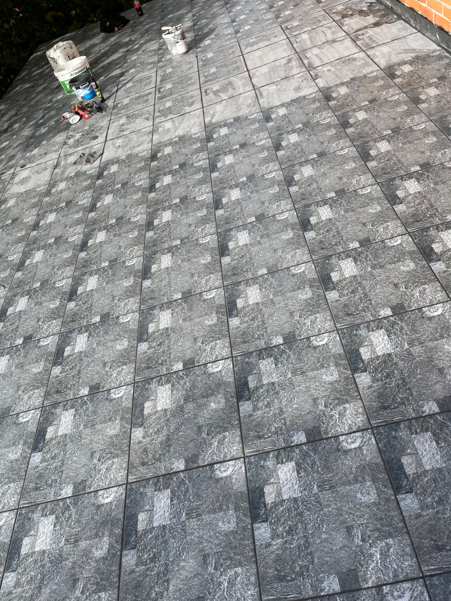 Ułożona nawierzchnia z szarych, kwadratowych płytek imitujących kamień, widoczne wiaderka z zaprawą i narzędzia do fugowania na krawędzi.