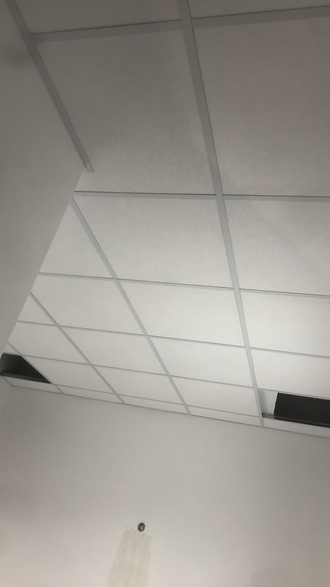 Sufit podwieszany z białymi panelami i metalową konstrukcją, widoczne niedokończone fragmenty i ślady po instalacji elektrycznej na ścianie.