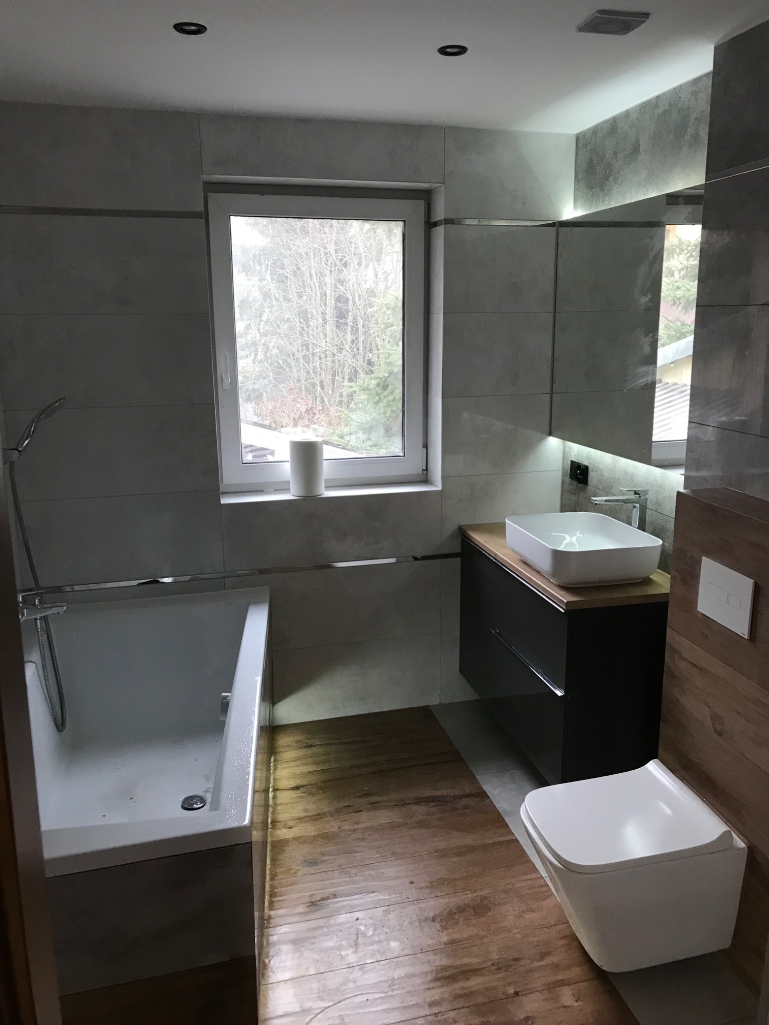 Nowoczesna łazienka z drewnianą podłogą, szarą glazurą, białą wanną i umywalką na szafce, oraz podwieszaną toaletą. Za oknem widok na drzewa.