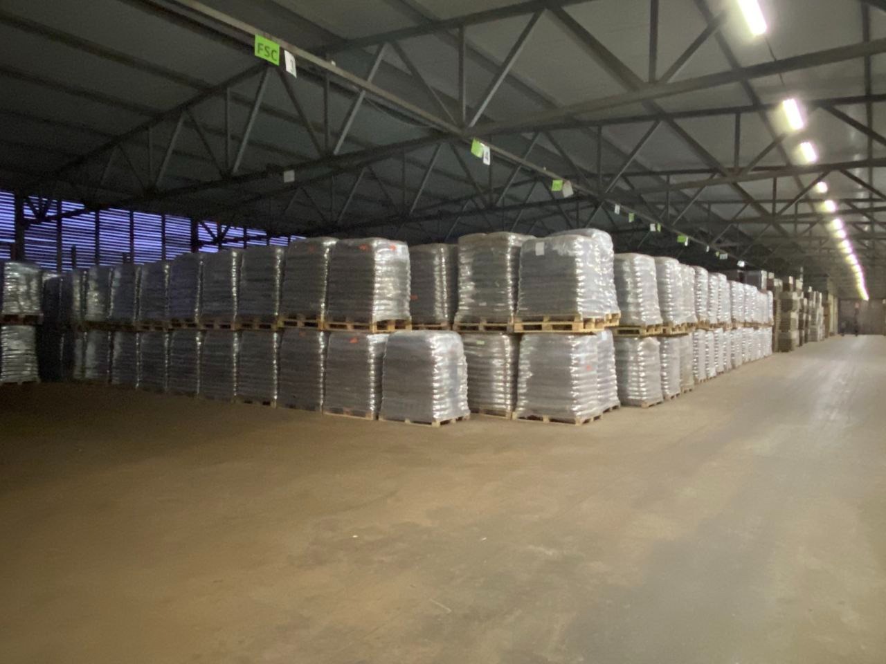Magazyn wypełniony paletami z workami opału, owiniętymi w przezroczystą folię, widoczne oznaczenia FSC na suficie.