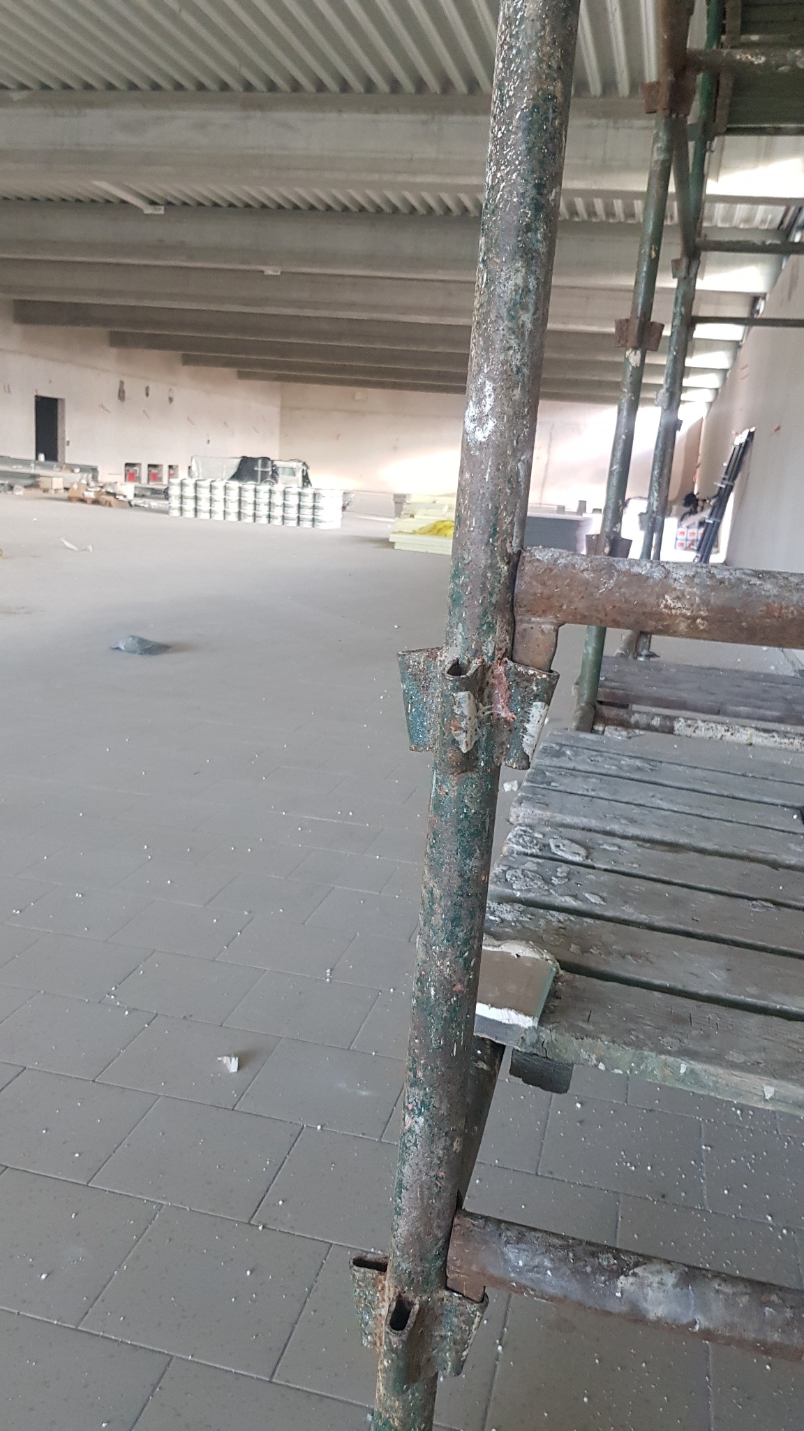 Metalowe rusztowanie w surowym wnętrzu, widoczne ślady użytkowania i farby, w tle przygotowane materiały budowlane, cegły i puszki z farbą.