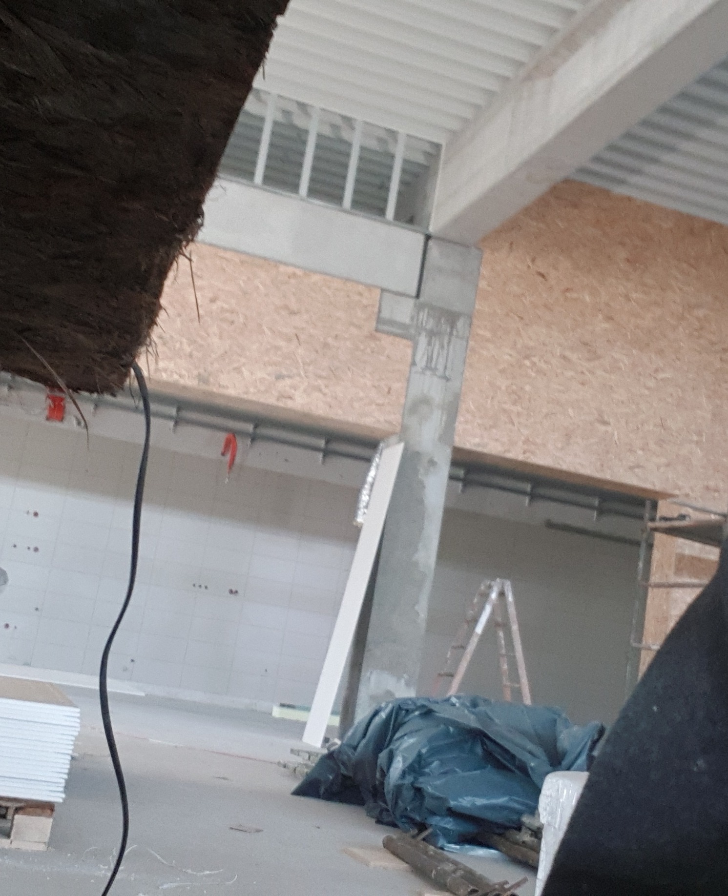 Wnętrze hali w Kartuzach w trakcie wykańczania: widoczna izolacja poddasza, płyty gipsowe, elementy konstrukcyjne i drabina.