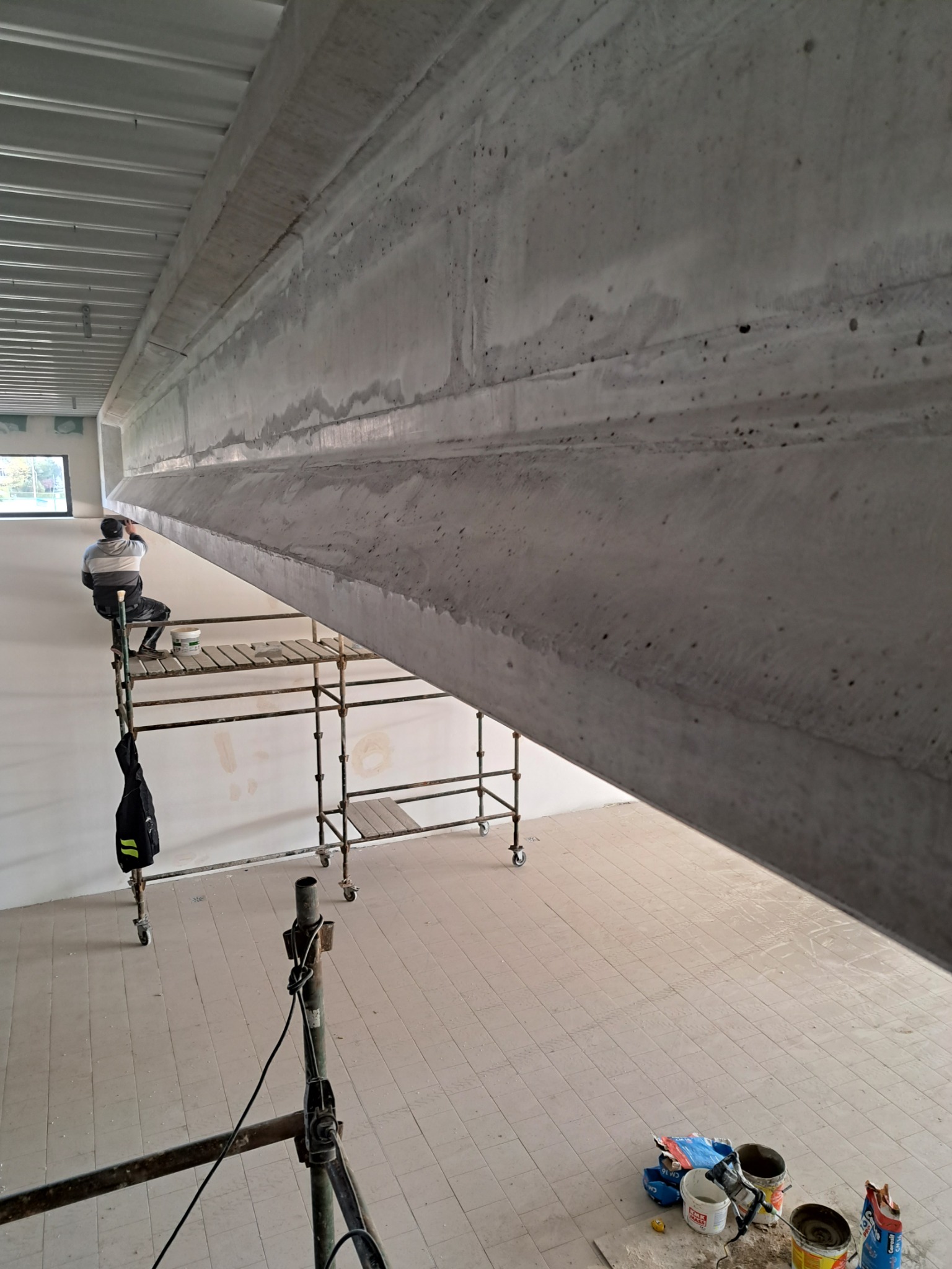 Pracownik na rusztowaniu wygładza betonową belkę pod sufitem w jasnym, przestronnym wnętrzu. Widoczne narzędzia malarskie i worki z zaprawą na podłodze.