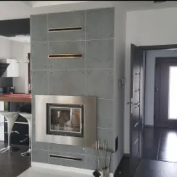 Nowoczesny salon z szarym kominkiem obłożonym płytkami, otwartą kuchnią z białymi meblami i wejściem do przedpokoju z ciemnymi drzwiami.