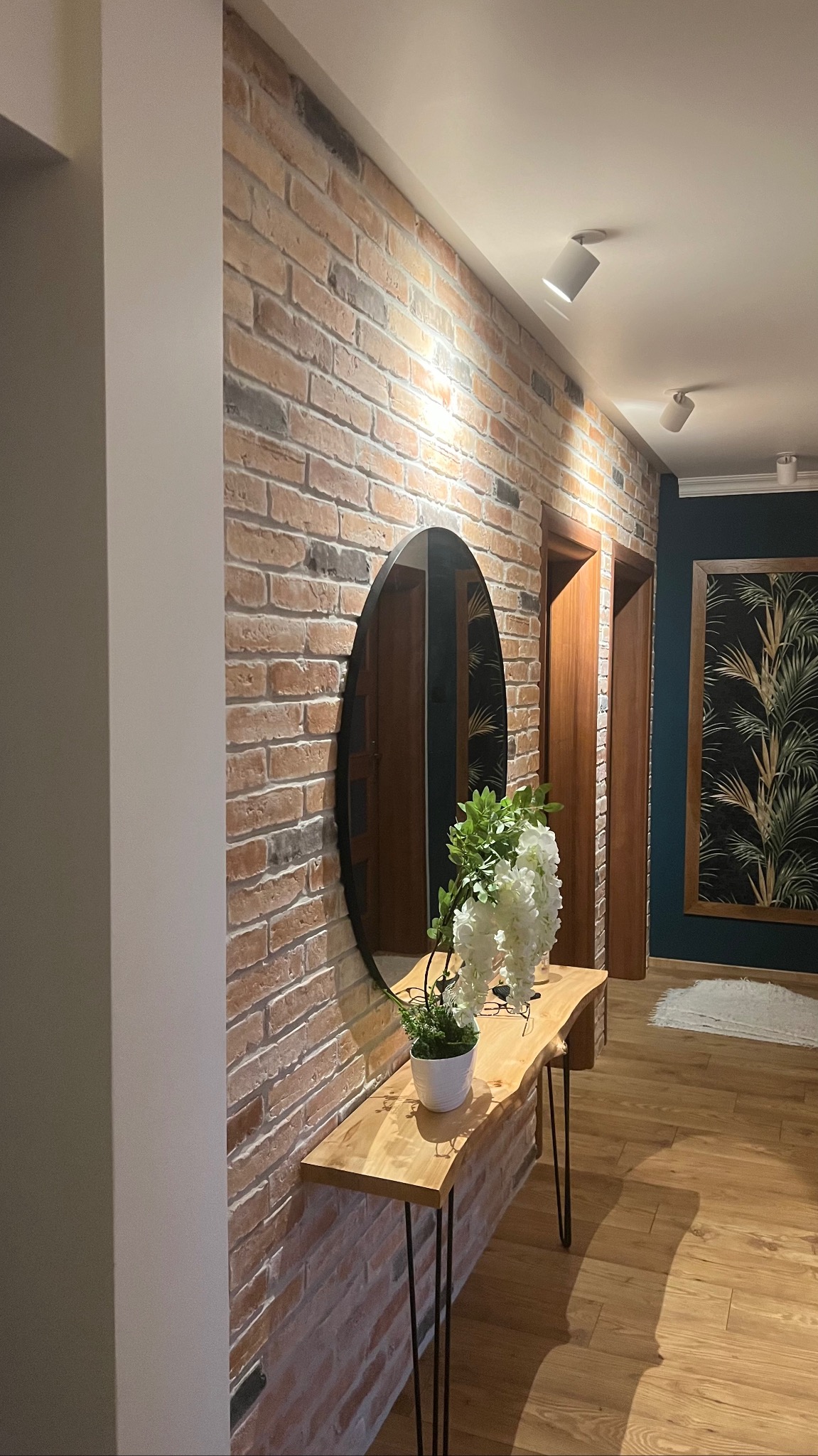 Wnętrze przedpokoju z ceglaną ścianą, owalnym lustrem, drewnianą konsolą z kwiatami i oświetleniem punktowym.