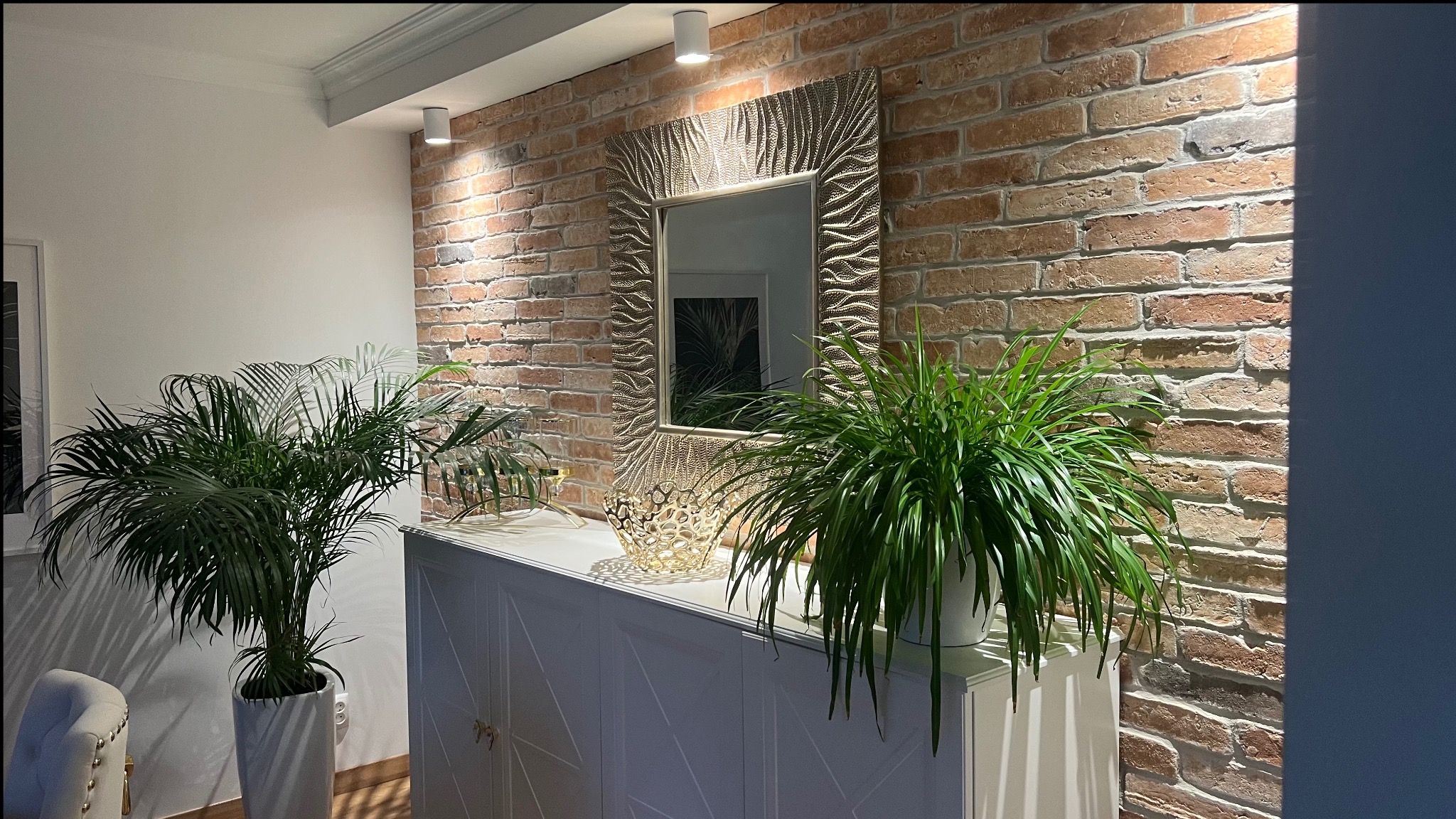 Udekorowane wnętrze: ceglana ściana z lustrem w ozdobnej ramie, biała komoda z geometrycznym wzorem, dwie rośliny doniczkowe i dekoracyjny lampion.