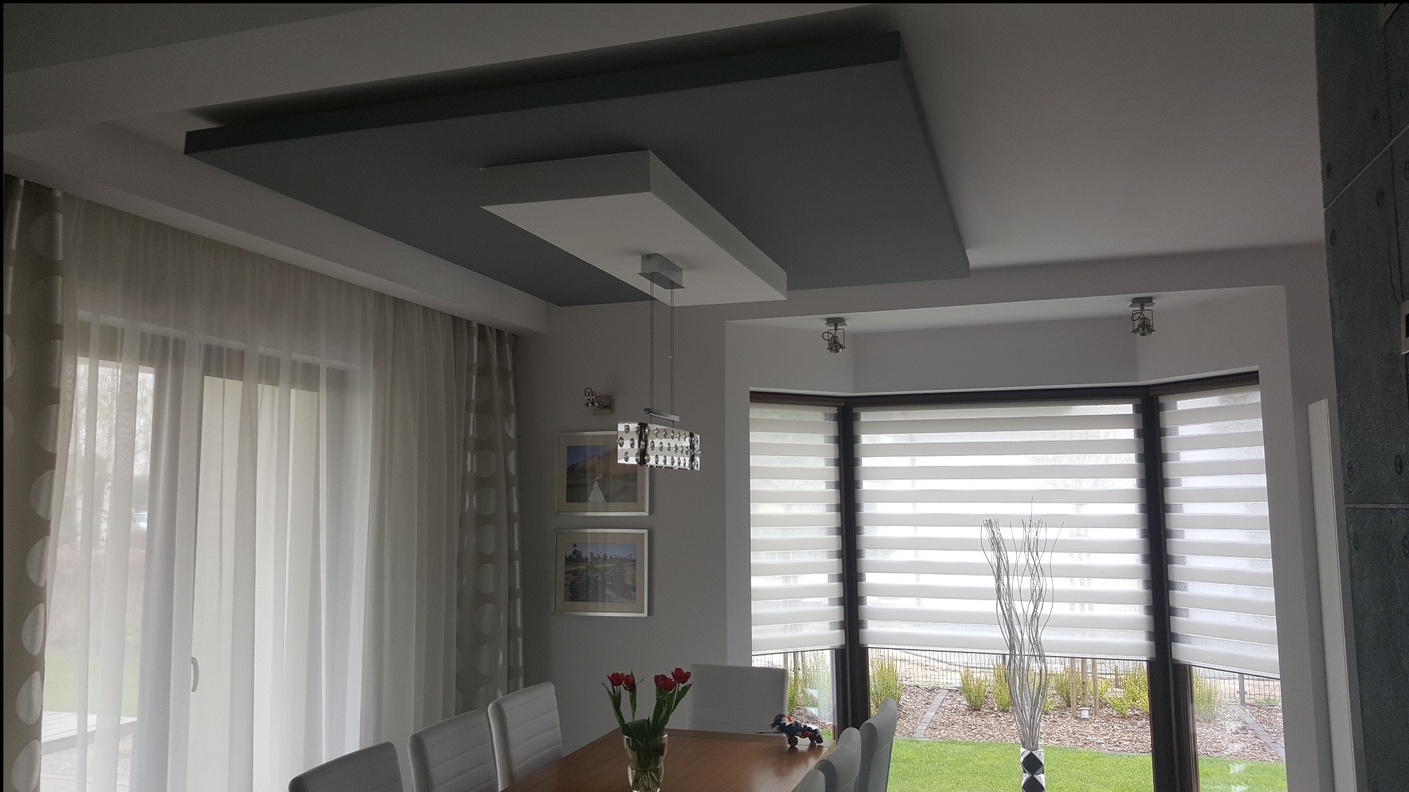 Nowoczesny salon z podwieszanym sufitem w odcieniach szarości i bieli, kryształowy żyrandol, okno z roletami dzień-noc i drewniany stół z czerwonymi tulipanami.