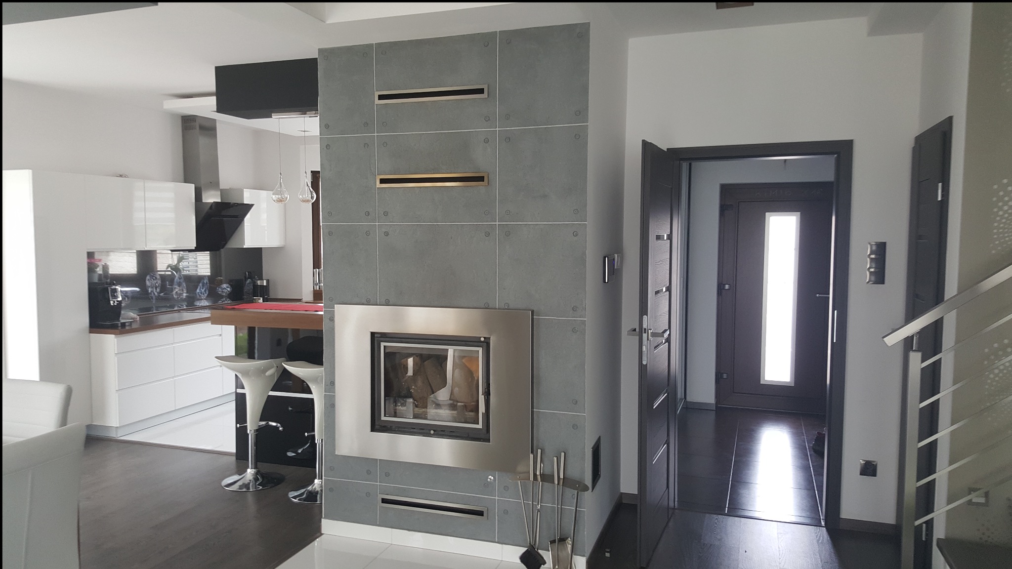 Nowoczesny salon z szarym kominkiem obłożonym płytkami, otwartą kuchnią z białymi meblami i wejściem do przedpokoju z ciemnymi drzwiami.
