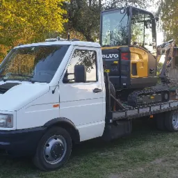 Biała laweta z żółtą mini koparką Volvo na platformie, zaparkowana na trawiastym terenie z drzewami i budynkiem w tle.